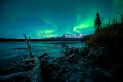 A wide-angle view showing a more diffused green aurora glow over snowy mountain peaks and a foreground river with logs.